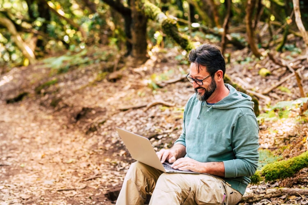 Ein Mann sitzt mit seinem Laptop in der Natur und schaut sich ein Lernvideo an.