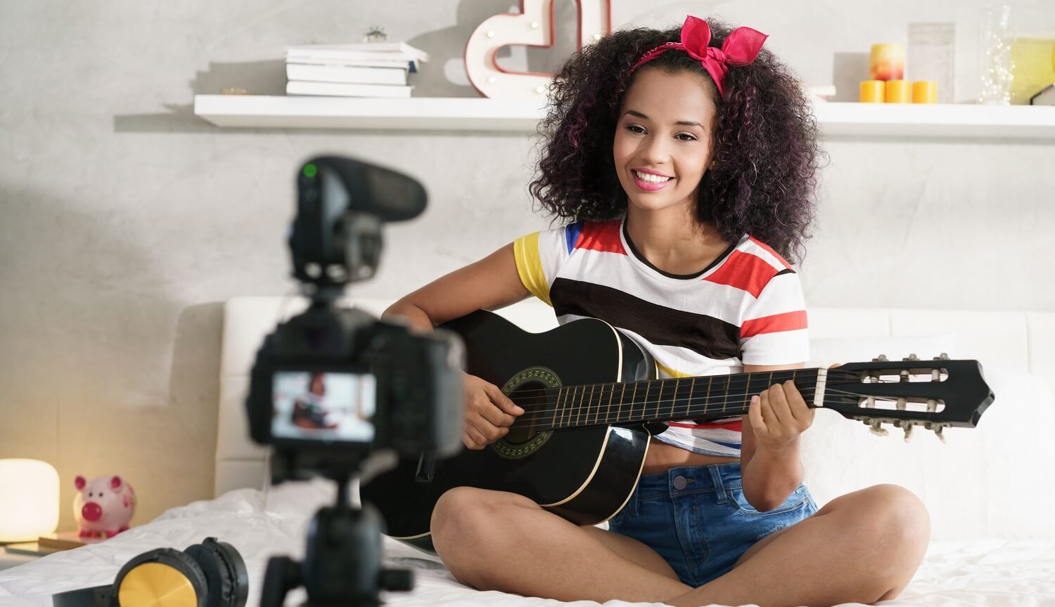 Eine Frau sitzt auf einem Bett und filmt ein Webvideo, in dem sie Gitarre spielt.