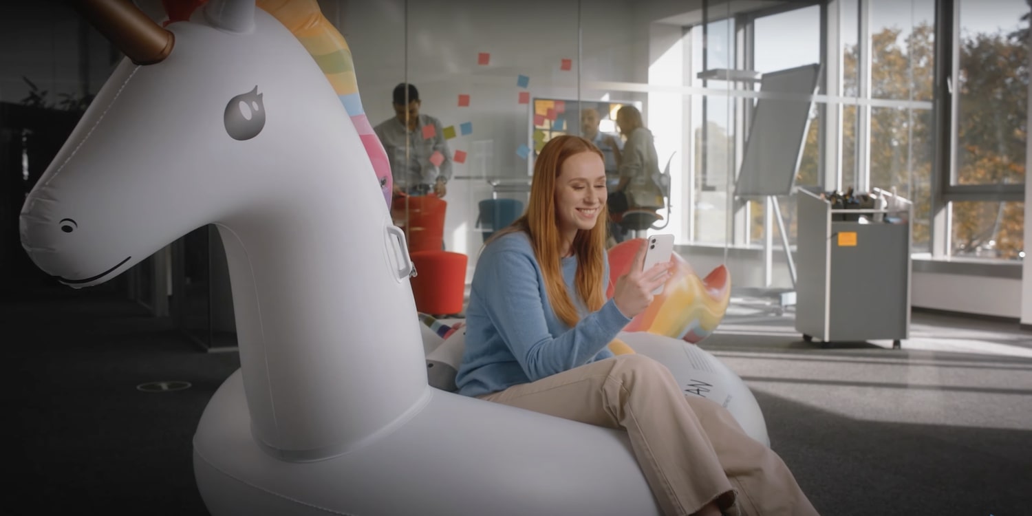 Eine Frau sitzt auf einer Einhorn-Luftmatratze und lächelt mit ihrem Handy in der Hand.