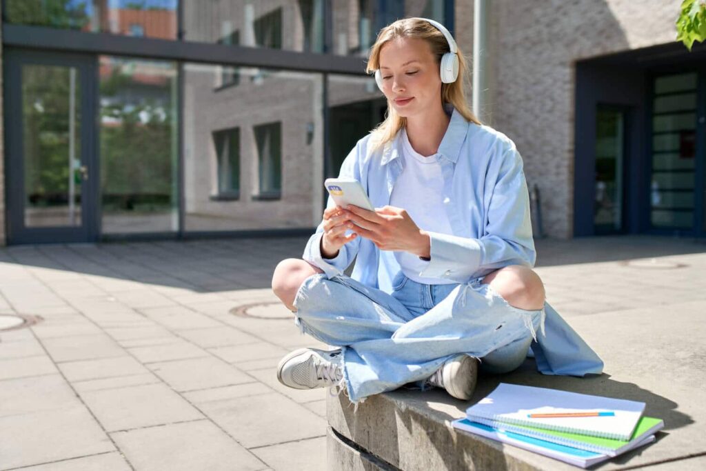Eine junge Frau sitzt vor ihrer Schule und hört während ihrer Mittagspause eine Podcast Episode.