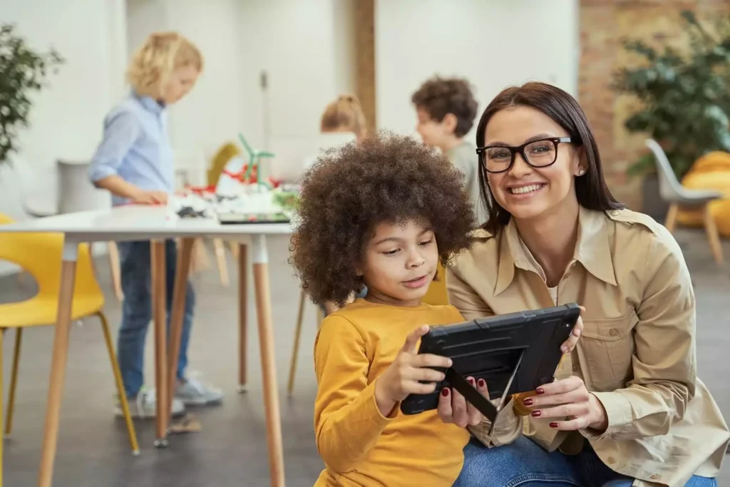Eine Lehrerin präsentiert ein Erklärvideo im Unterricht, während Schüler im Hintergrund an einem Tisch arbeiten.